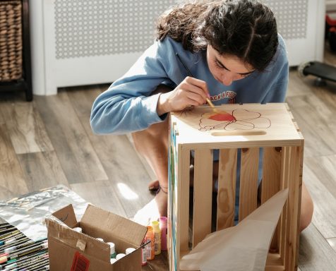Crafty!! A girl painting a wooden crate on a wooden floor with craft materials around her.